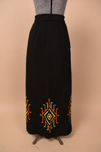 Load image into Gallery viewer, Vintage 1960's black column maxi skirt is shown from the front. This skirt has a red, green, and yellow embroidered design at the bottom hem.