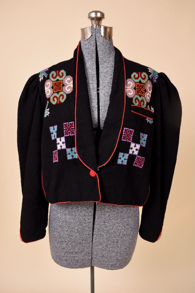 Vintage 1980's cropped black blazer is shown from the front. This embroidered bolero jacket has colorful folksy cross stitch embroidery on the front. 