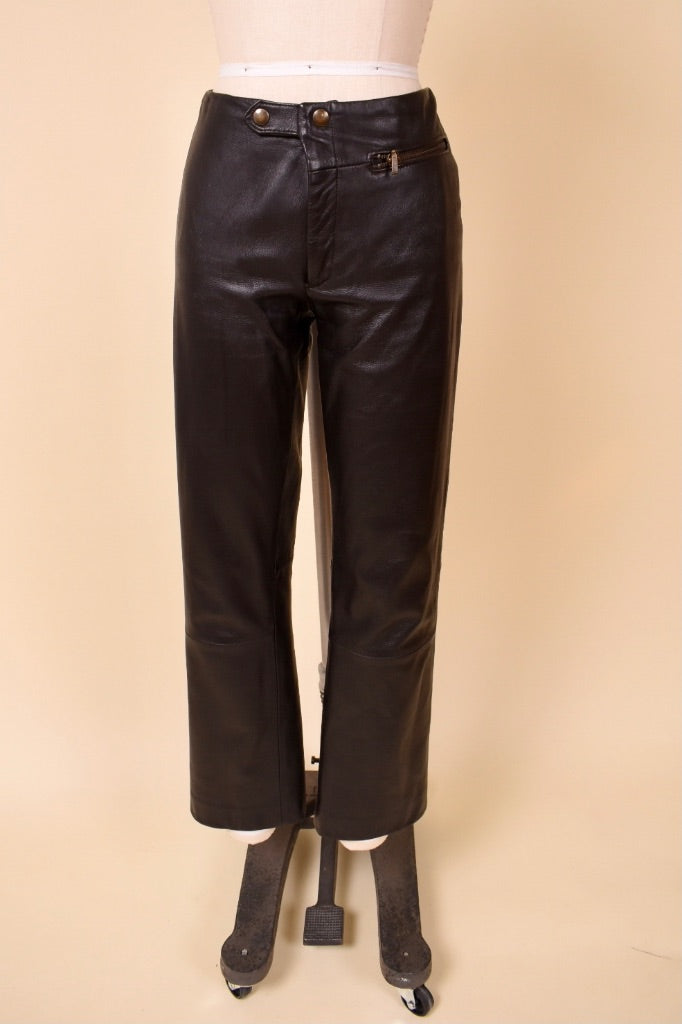 Vintage Y2K brown leather low rise moto pants are shown from the front. These pants have gold buttons and zippers at the waist. 