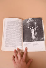 Load image into Gallery viewer, A hand holds open the book. The left page has text. The right page has a black and white photograph. A person has their arms splayed out in the photo.
