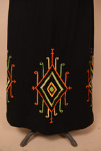 Load image into Gallery viewer, Vintage 1960s black cotton maxi skirt is shown in close up. This skirt has an artsy red, green, and yellow diamond shaped design at the bottom hem.
