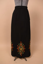 Load image into Gallery viewer, Vintage 1960&#39;s black column maxi skirt is shown from the front. This skirt has a red, green, and yellow embroidered design at the bottom hem. 
